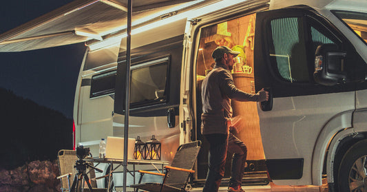 A man enters his RV camper through a mesh screen door. Warm light glows from inside and the side of the vehicle.