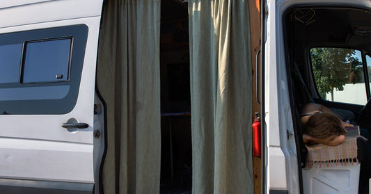 A side view of a van with open doors and beige curtains hanging from the entrance. A traveler rests in the front seat.