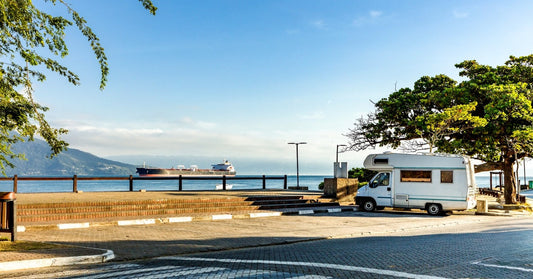 A view of a coastline with a large cargo ship entering the harbor. An RV camper is parked next to a tree.