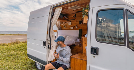 A man wearing a blue baseball cap sits on the floor of his open van while holding a cup and staring at the road.