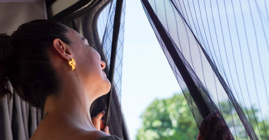 A woman slightly opens the insulated curtains of her camper van and gazes out at a sunny environment.