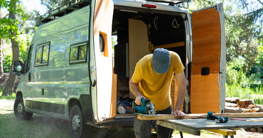A man saws a piece of wood at a workbench outside near his camper van. The rear doors of the vehicle are open.