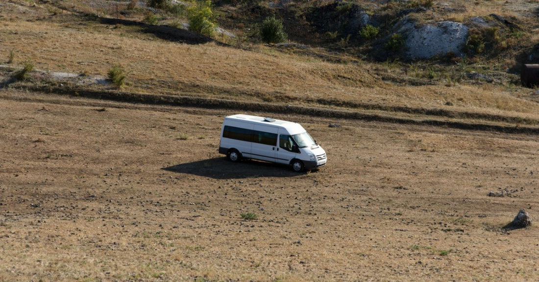 A white camper van parked on dry rocky land with sparse vegetation and distant hills on a bright sunny day.