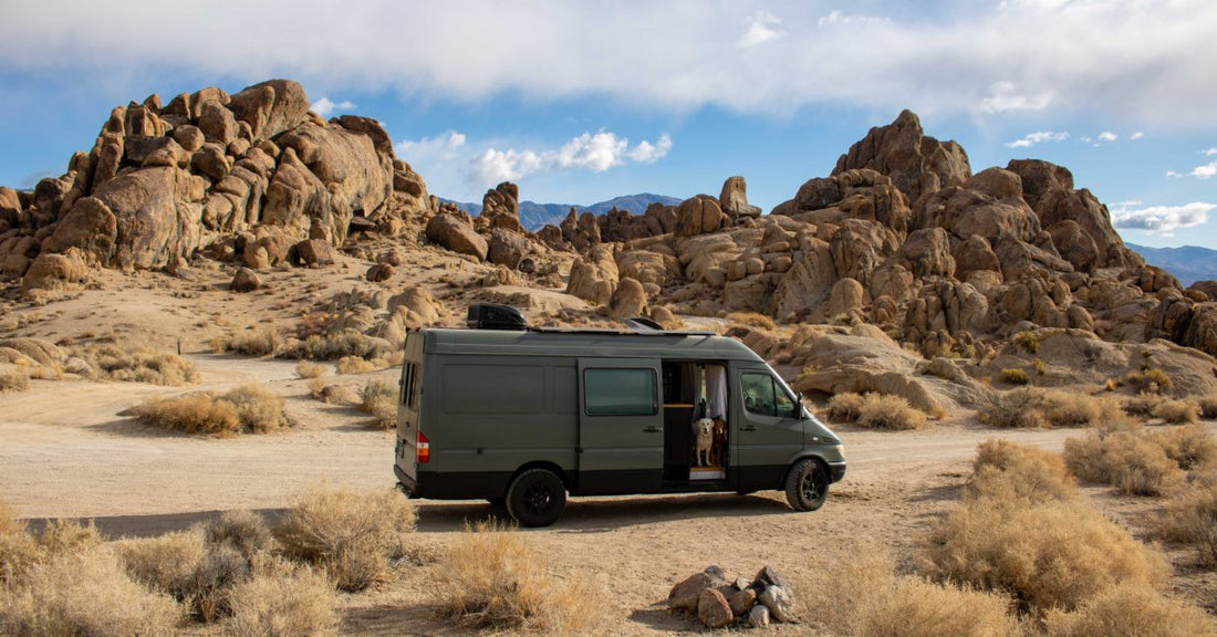 A dark camper van parked in a desert near large rock formations, dry brush, and mountains under a cloudy blue sky.