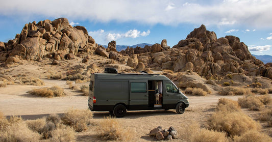 A dark camper van parked in a desert near large rock formations, dry brush, and mountains under a cloudy blue sky.