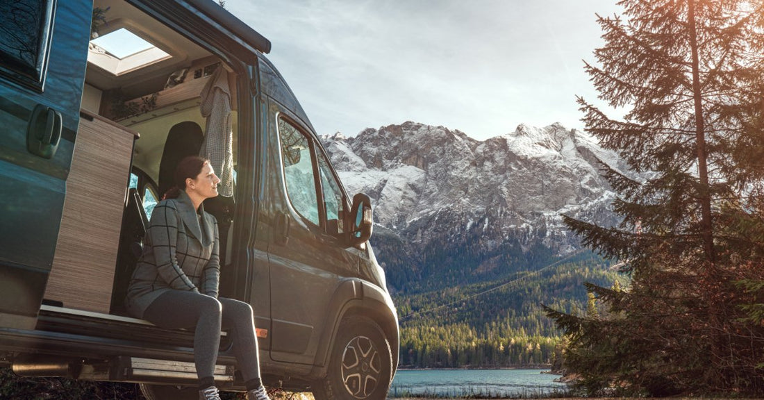 A low-angle view of a woman sitting on the open side of a camper van, with mountains and sky behind her.