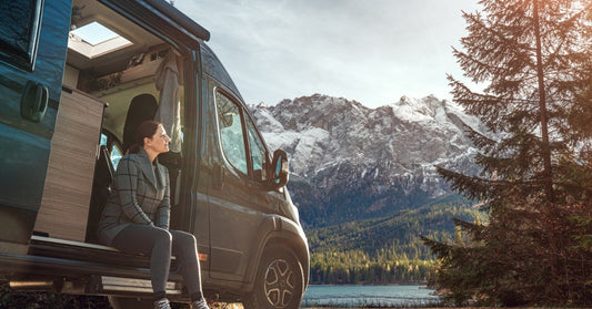 A low-angle view of a woman sitting on the open side of a camper van, with mountains and sky behind her.