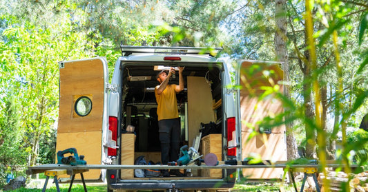A man installing a ceiling light inside an open camper van surrounded by trees, tools, and wooden doors.