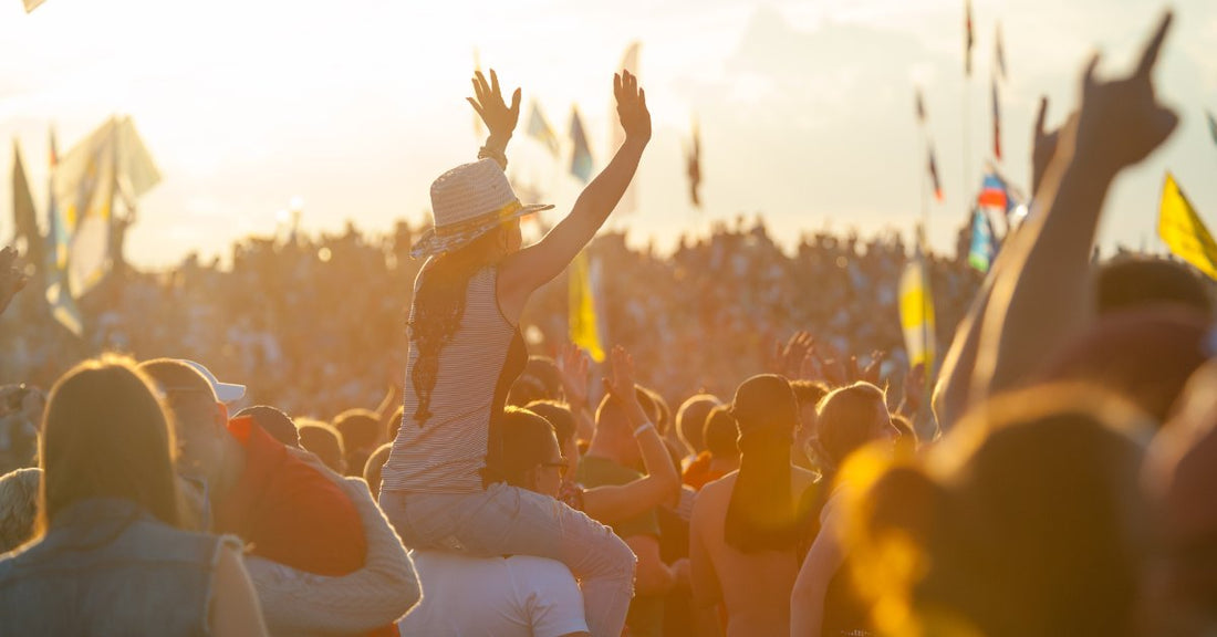 A crowd gathered outdoors at a music festival. They have their hands raised, with stage lights in the distance.