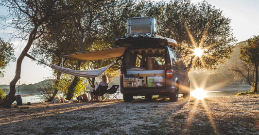 A dark camper van is parked by the trees, with a hammock near the water as sunlight filters through the trees at the camp.