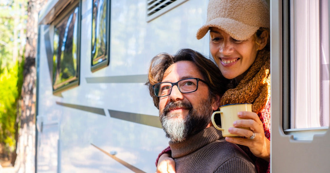 A close-up of two adults standing at a camper van doorway with their mugs, and trees surrounding the vehicle.