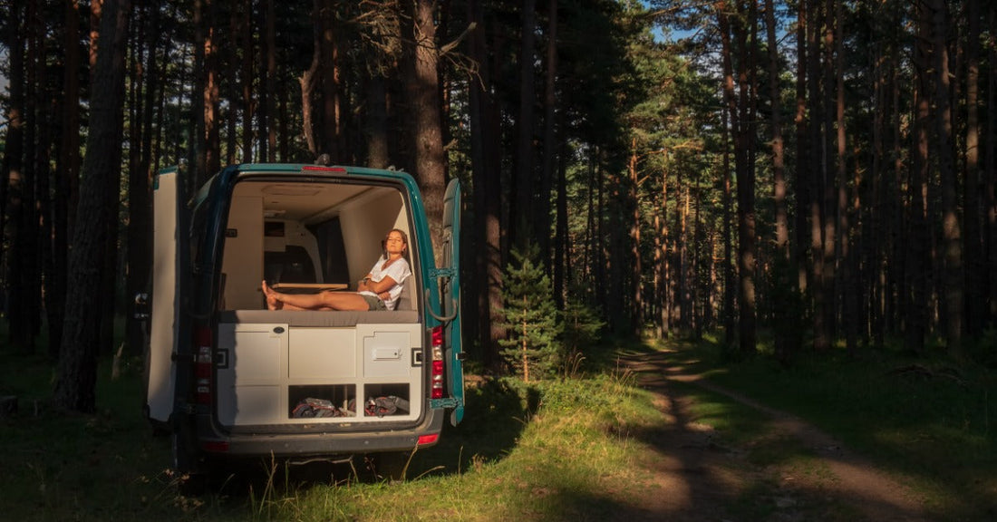 A close-up of a person sitting in the open doorway of a parked camper van with a vast forest in the background.