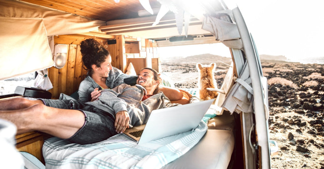 A man and a woman are lying on a bed inside a camper van, with a small dog looking out at the remote landscape.