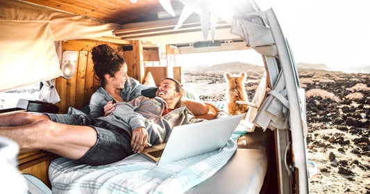A man and a woman are lying on a bed inside a camper van, with a small dog looking out at the remote landscape.