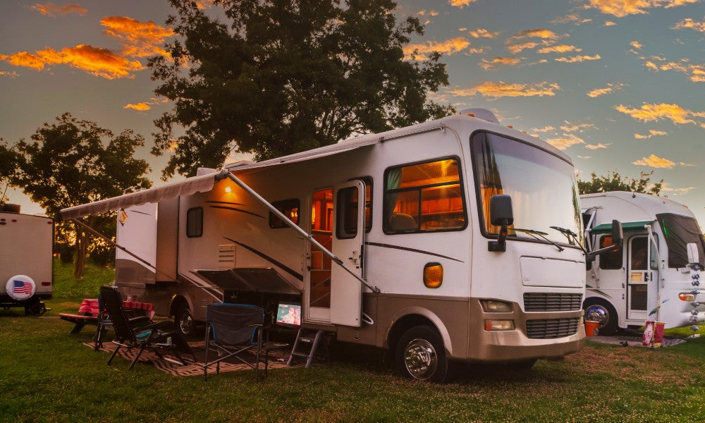 An RV parked at a campground with chairs and a picnic table underneath the extended awning. The lights are on in the RV.