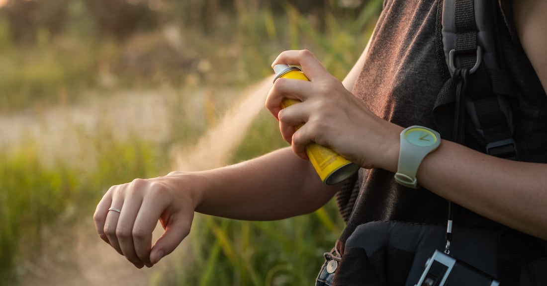 A woman is standing outside in a grassy area. She is spraying a yellow can of bug spray on her right arm.