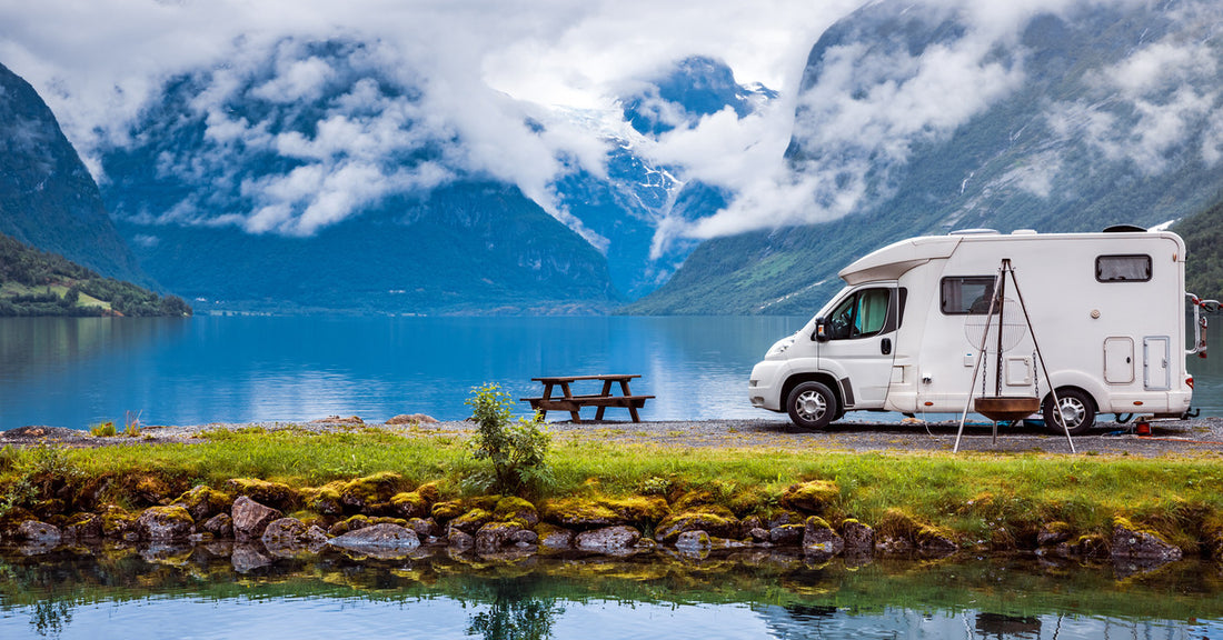 A white RV parked next to a lake surrounded by mountains. There are clouds around the mountains and a picnic table next to the RV.