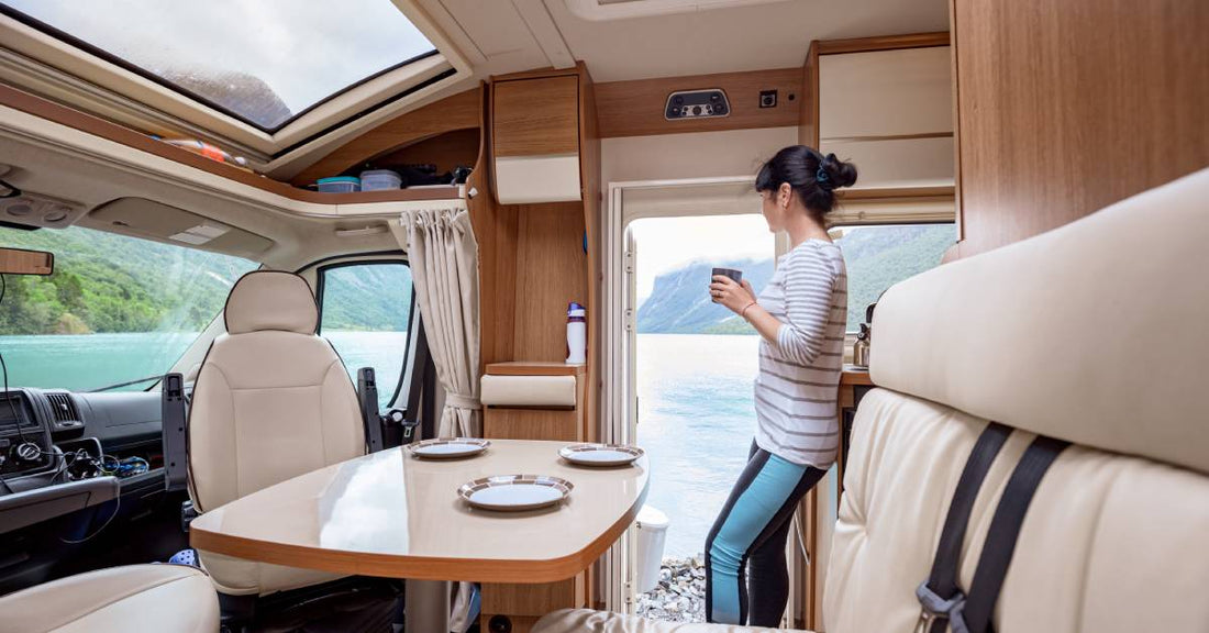 A woman standing inside an RV and holding a cup of coffee. The door is open and she is looking out at a body of water.