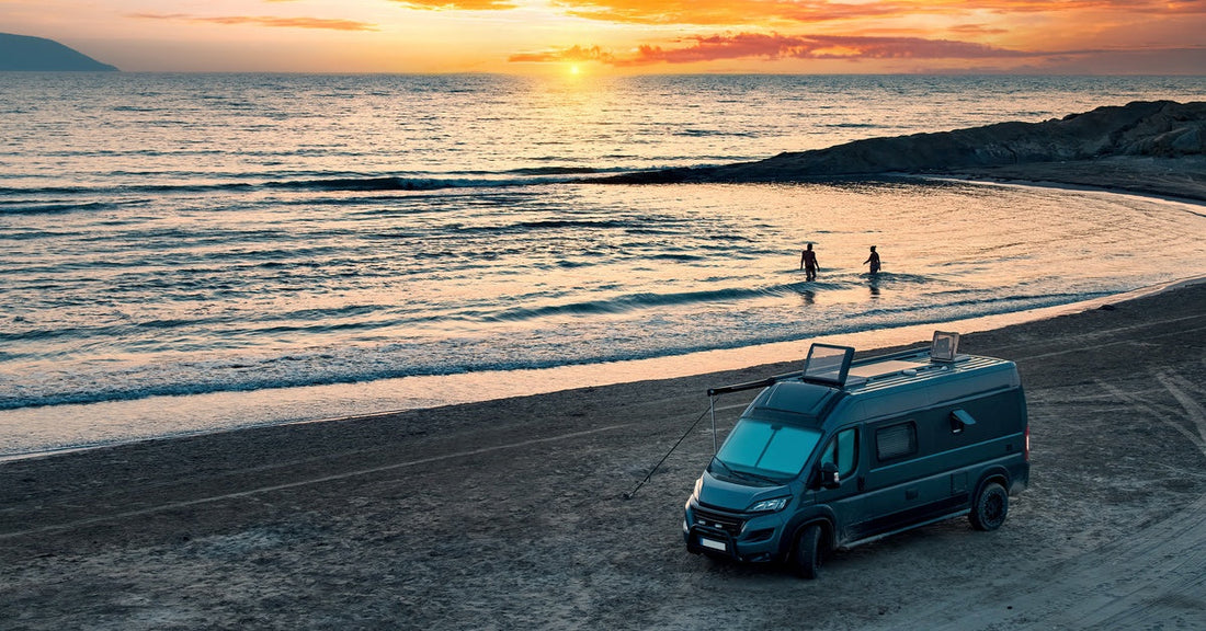 An aerial view of a camper van on a beach against a beautiful sunset. Two individuals bathe in the sea.