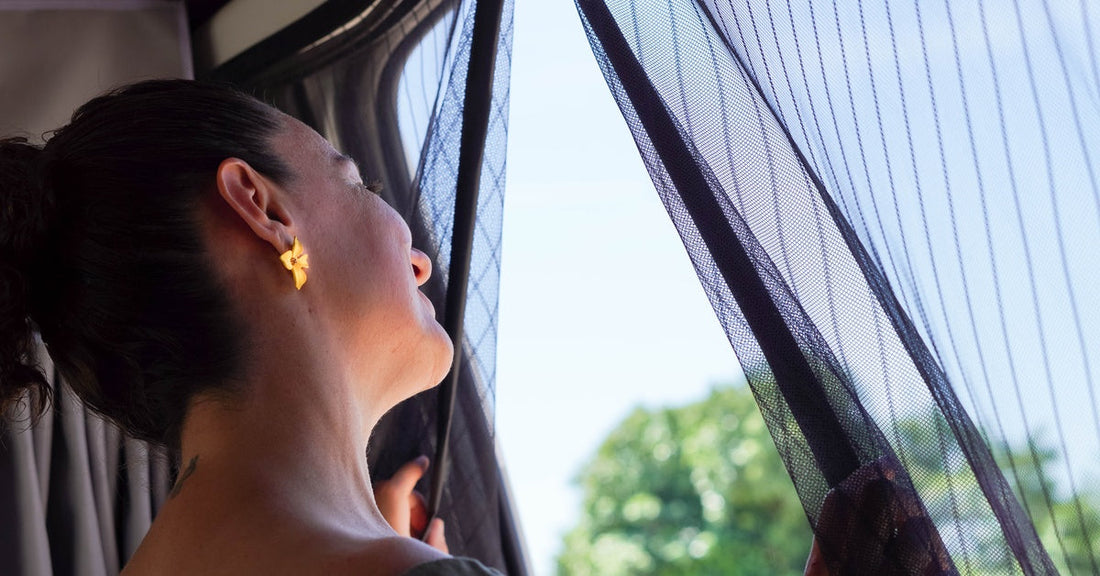 A woman slightly opens the insulated curtains of her camper van and gazes out at a sunny environment.