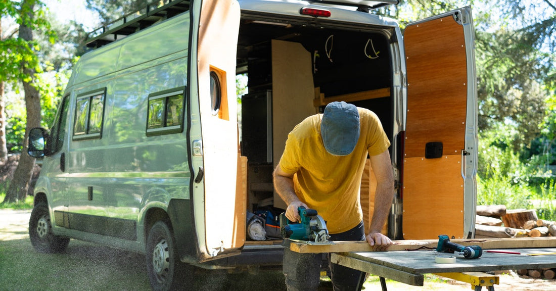 A man saws a piece of wood at a workbench outside near his camper van. The rear doors of the vehicle are open.