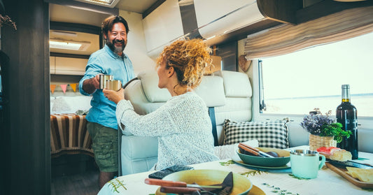 A man stands by a doorframe, raising his glass to a woman seated at a table set with plates and a bottle.