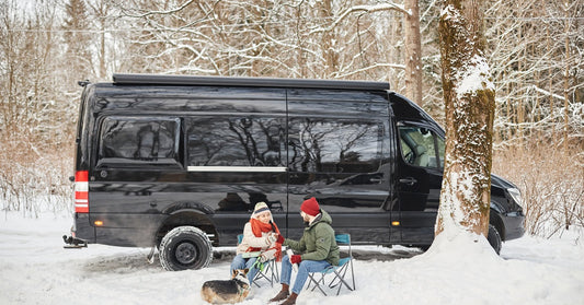 A man and woman, wearing their winter coats, sit with a dog in front of a black camper van parked in a snowy forest.