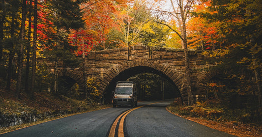 A van drives under a stone bridge on a curving road, surrounded by vibrant autumn trees in red, orange, and yellow.