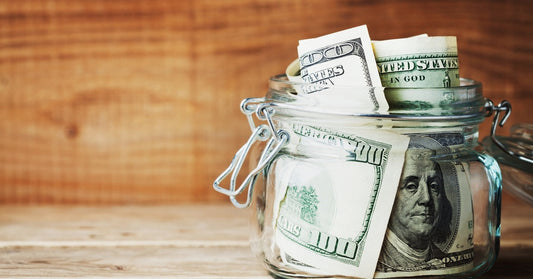 A small, clear jar on the corner of a wooden surface is open and filled with several hundred-dollar bills.