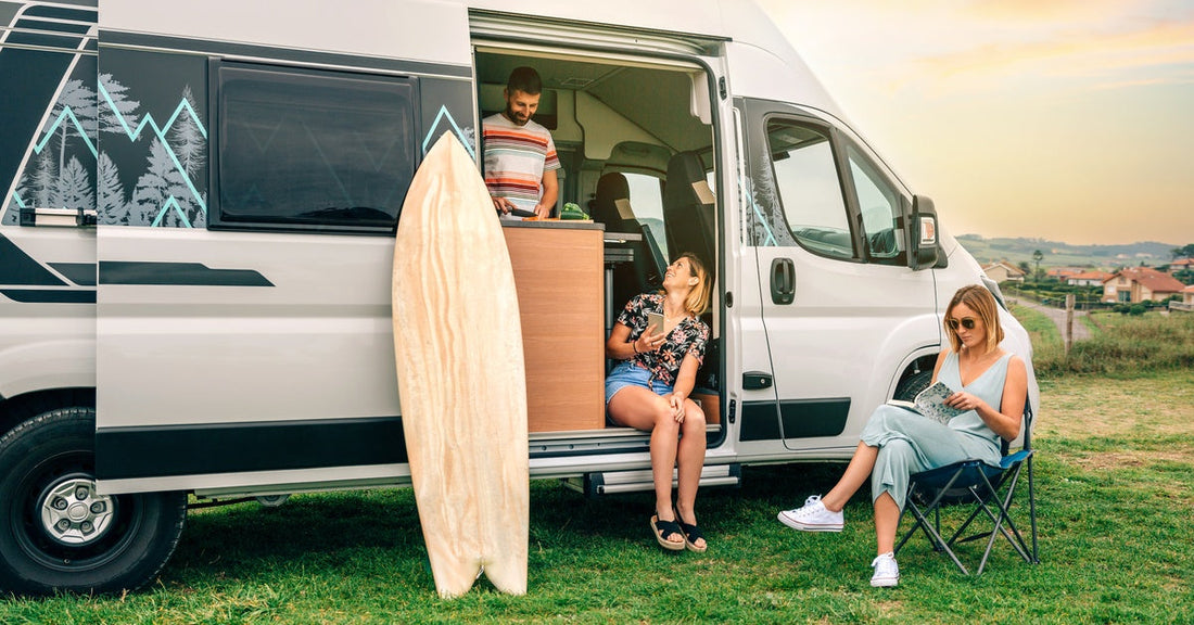 Two women sit by a parked camper van in a green field, while a man stands inside the vehicle next to wooden furniture.