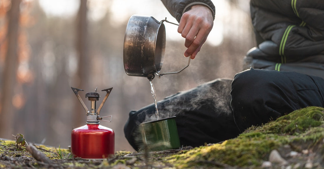 A camper pours hot water from a metal pot into a green cup, with steam rising, near a mini stove on the floor.