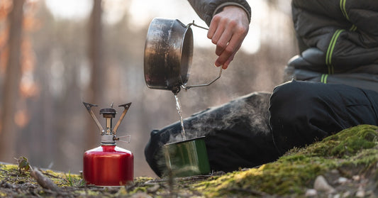 A camper pours hot water from a metal pot into a green cup, with steam rising, near a mini stove on the floor.