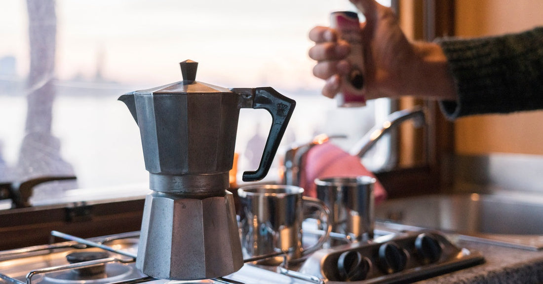 Moka pot on a gas stove with a blue flame, a hand holding a lighter, and a blurred window in the background.