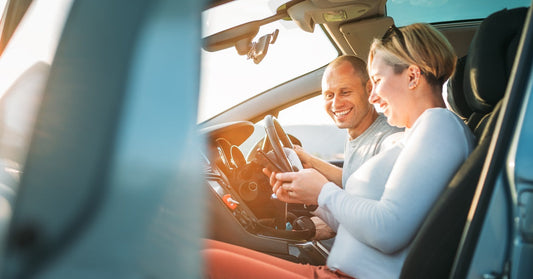 A man and woman sit in a sunlit car, sharing a moment of joy and leisure as they look at a smartphone together.
