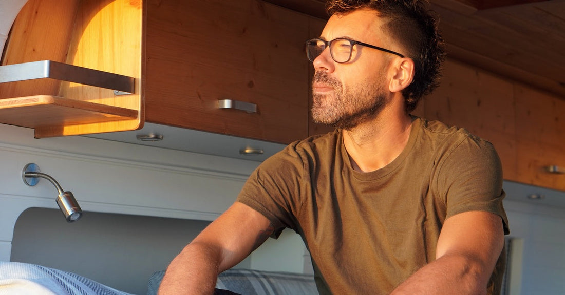 A Millennial man wearing glasses sits on a bed beneath cabinets, gazing outward as soft, warm light illuminates him.