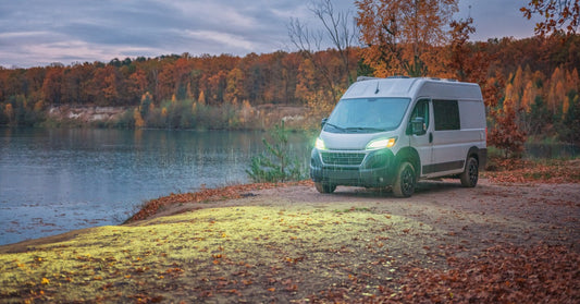 A wide shot of a parked gray campervan near the shore, surrounded by trees with orange leaves on a rainy day.