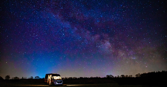 A wide shot of a parked campervan glowing with light from within, sitting in a dark field beneath a star-filled sky.