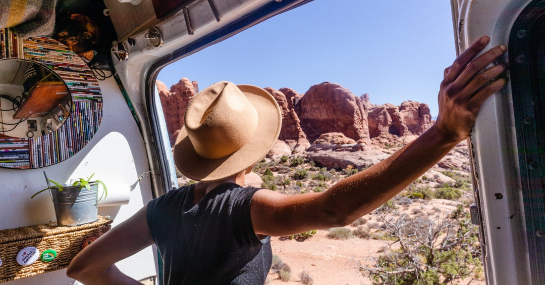 A person wearing a tan cowboy hat leans out of a camper van door, looking out at red rock desert formations.