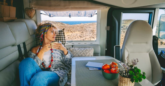 A woman relaxing inside a camper van, holding a mug and looking out at a coastal landscape through the side window.