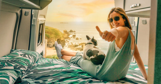 A young woman wearing sunglasses is sitting on a camper van bed with her dog, watching a colorful sunset outdoors.