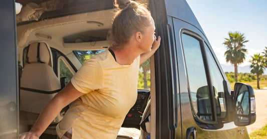 A woman leaning out of a camper van window near palm trees on a sunny day with blue sky in the background.