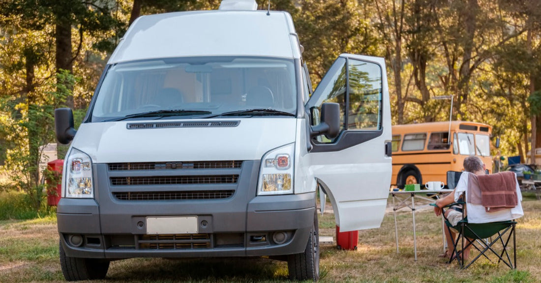 A person sits next to a parked camper van with the driver's door open. In the background, a school bus is visible.