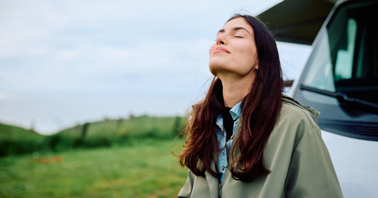 A woman with her eyes closed and her head pointed at the sky stands beside a camper van near trees and grass.