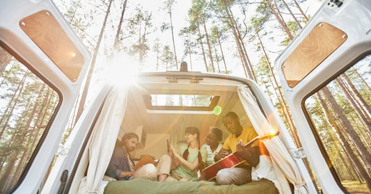 A low-angle view of four adults sitting in an open camper van in a forest, with one person playing the guitar.